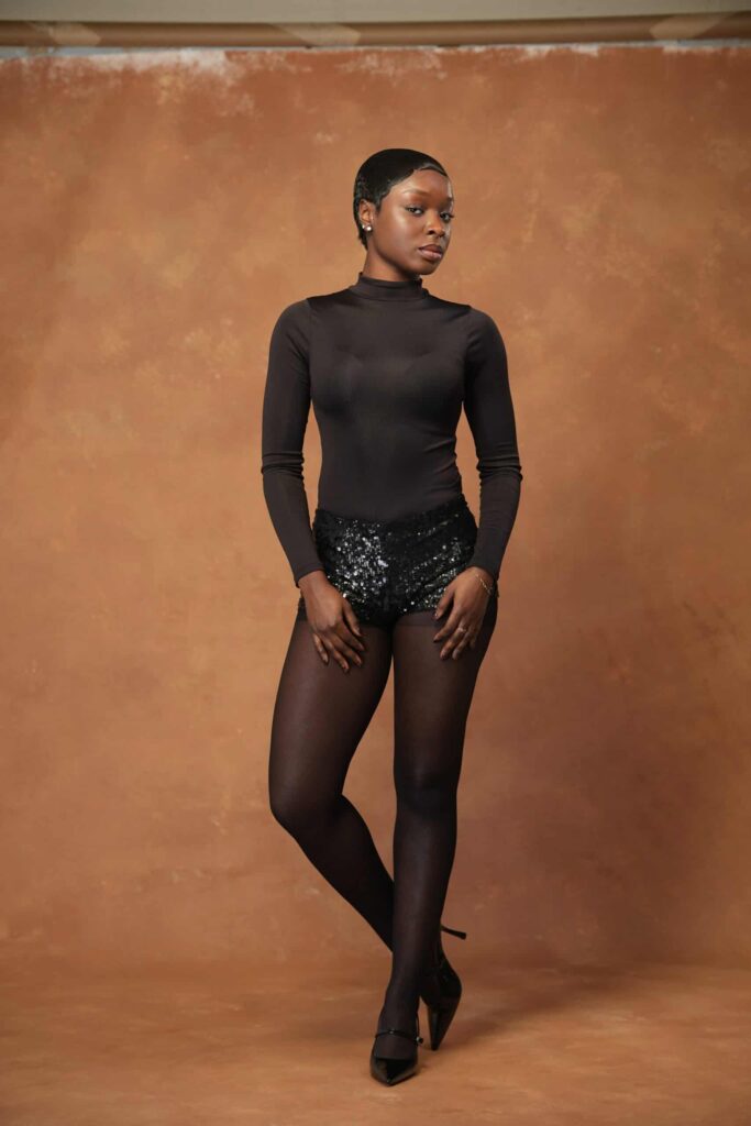 Woman in a sheer black bodysuit posing against a warm brown painted backdrop